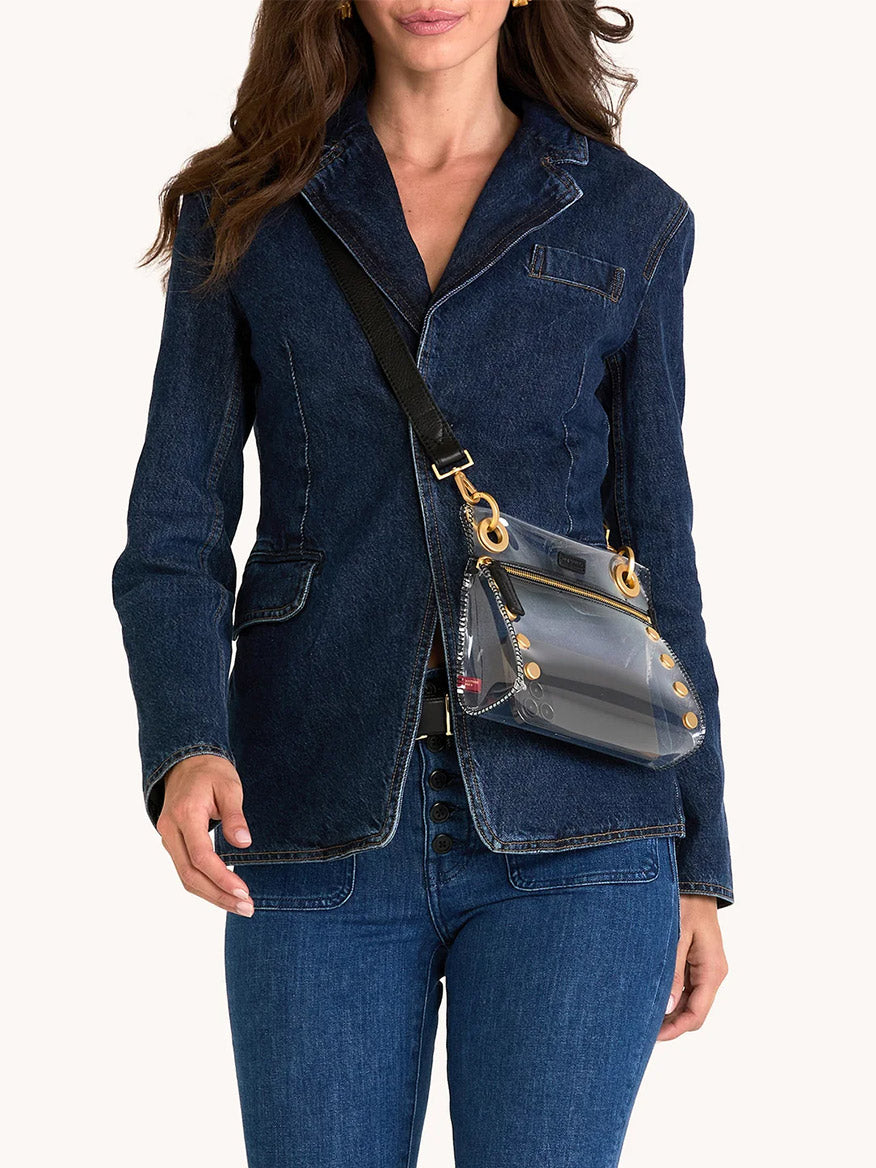 A person in a dark denim blazer and jeans carries the Hammitt Los Angeles Tony Small Crossbody Bag in Clear Black/Brushed Gold, featuring an adjustable strap, shown from mid-thigh up against a plain background.