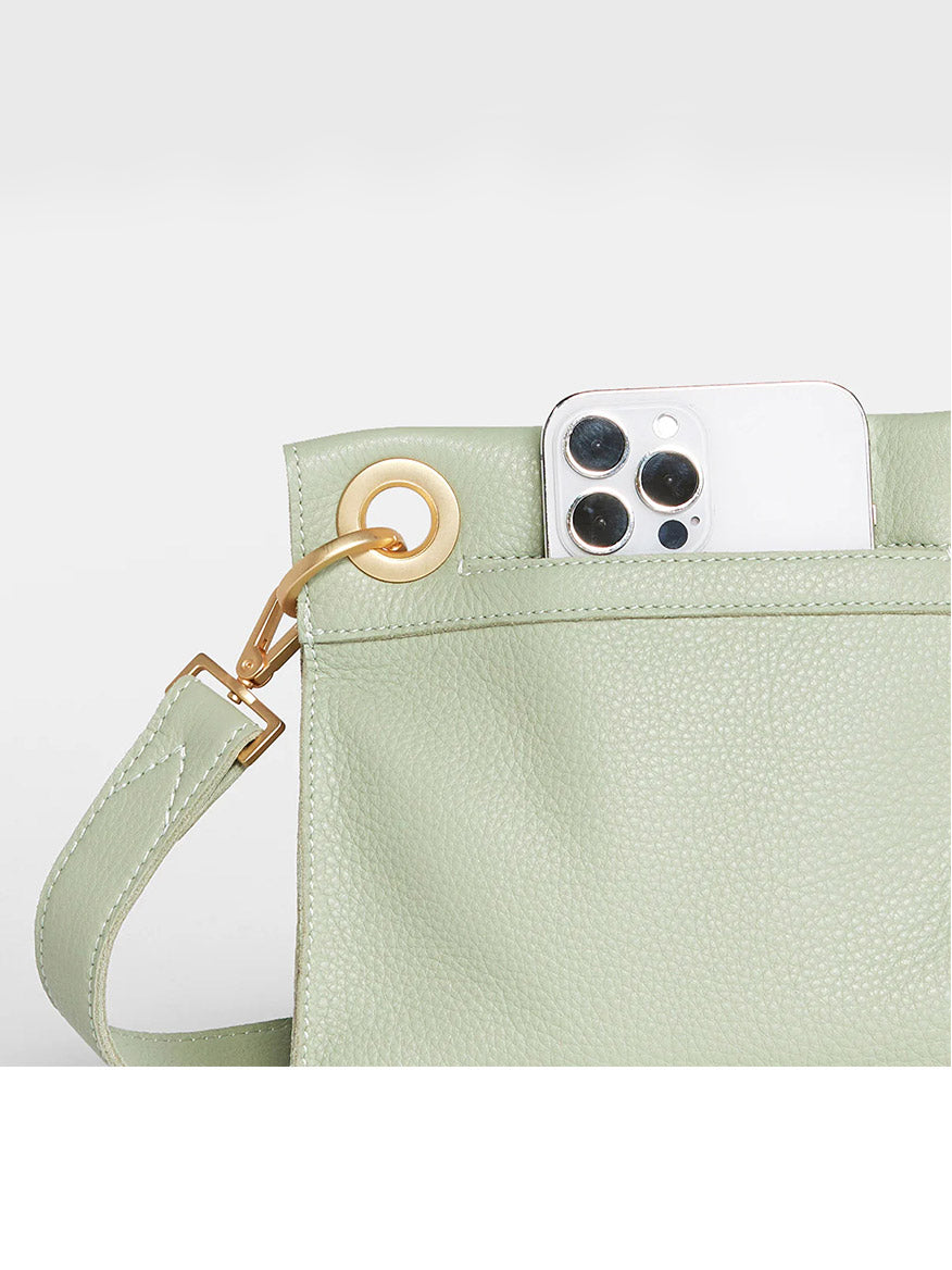 A smartphone with a triple-lens camera peeks out from the pocket of a Hammitt Los Angeles Tony Small Crossbody Bag in Cypress Sage with a gold clasp.