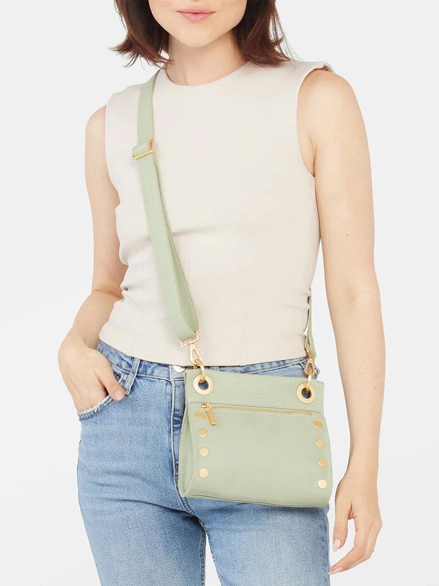 Woman in a sleeveless top and jeans, holding a Hammitt Los Angeles Tony Small Crossbody Bag in Cypress Sage with stud details.