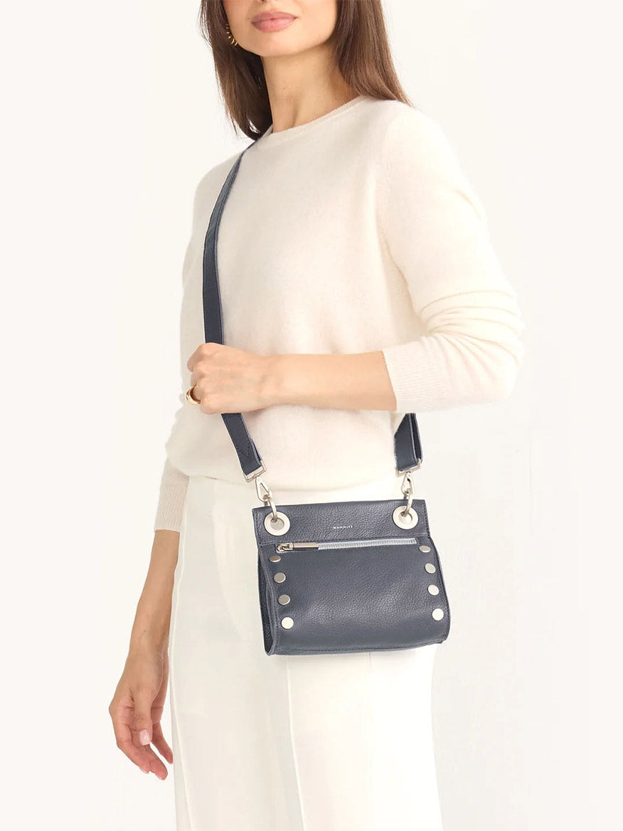 A woman in a white sweater and pants is holding a Hammitt Los Angeles Tony Small Crossbody Bag in Jazz Blues, featuring metal accents and an adjustable strap.