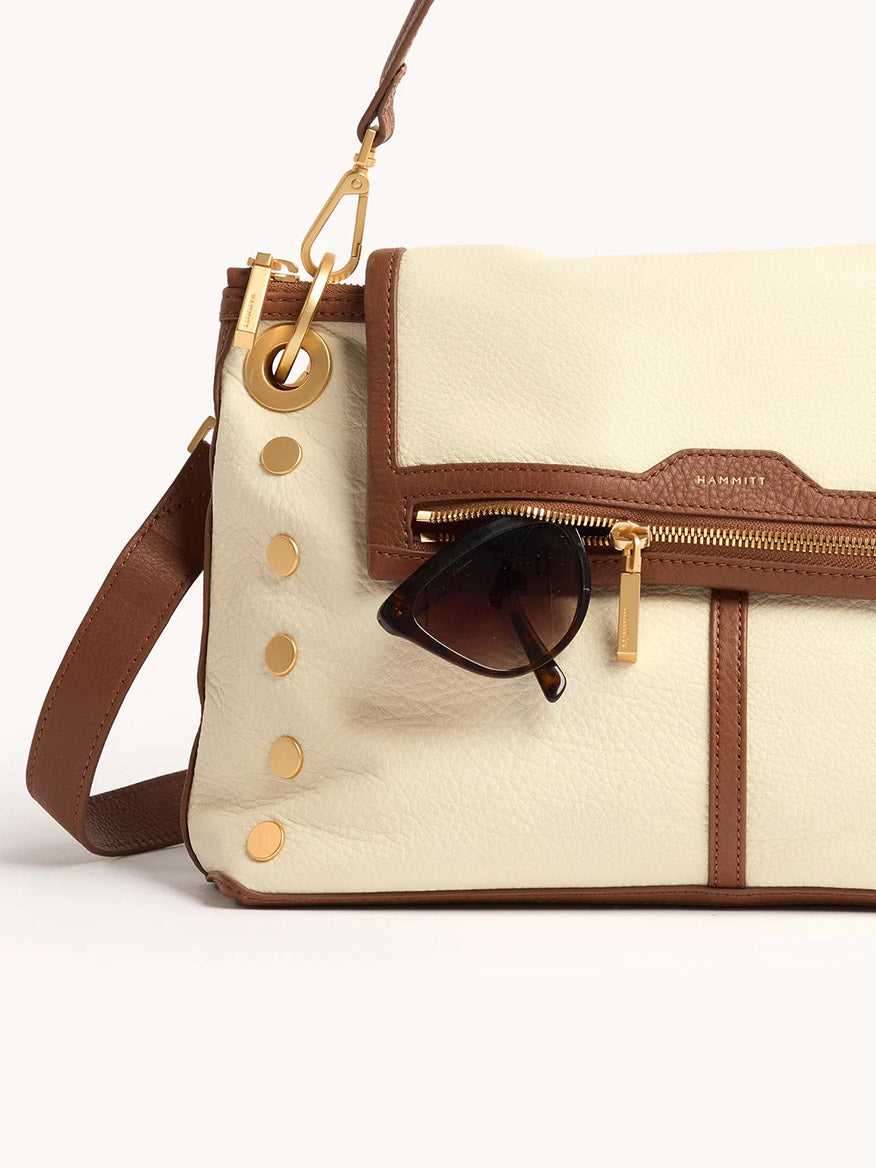 The Hammitt Los Angeles VIP Grand in Courtyard Cream is a cream and brown leather crossbody with gold hardware, inspired by signature styles, featuring a pair of sunglasses hooked onto its front pocket.