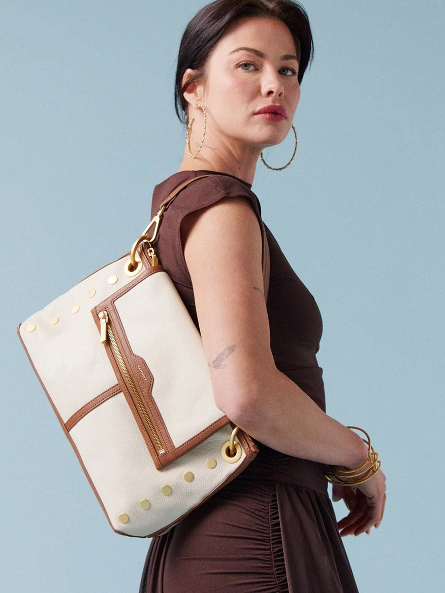 A woman in a brown dress carries the Hammitt Los Angeles VIP Grand in Courtyard Cream crossbody bag with gold accents over her shoulder, set against a light blue background.