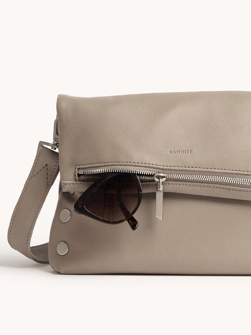 A Hammitt Los Angeles VIP Medium Clutch in Pewter Pebble leather with a silver zipper, shown with brown sunglasses partially tucked into its front pocket.