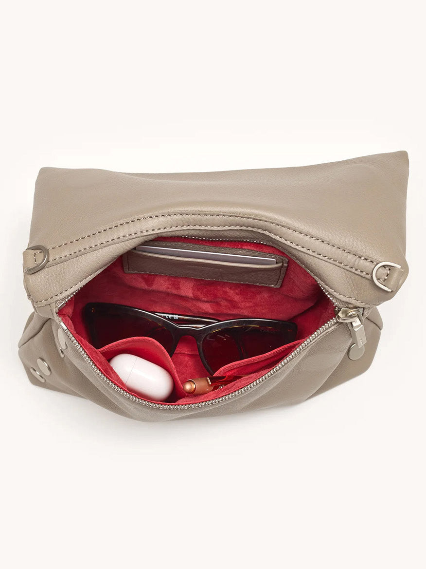 A Hammitt Los Angeles VIP Medium Clutch in Pewter Pebble is unzipped to reveal sunglasses, wireless earbuds, a lipstick, and a rectangular item inside its red interior.