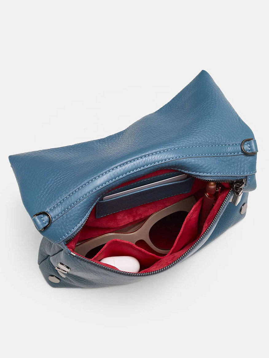 The Hammitt Los Angeles VIP Medium Clutch in Curated Blue is open, revealing a red interior with sunglasses, a phone, and a white round object inside. With its removable crossbody strap, this clutch effortlessly fuses style and function for any occasion.