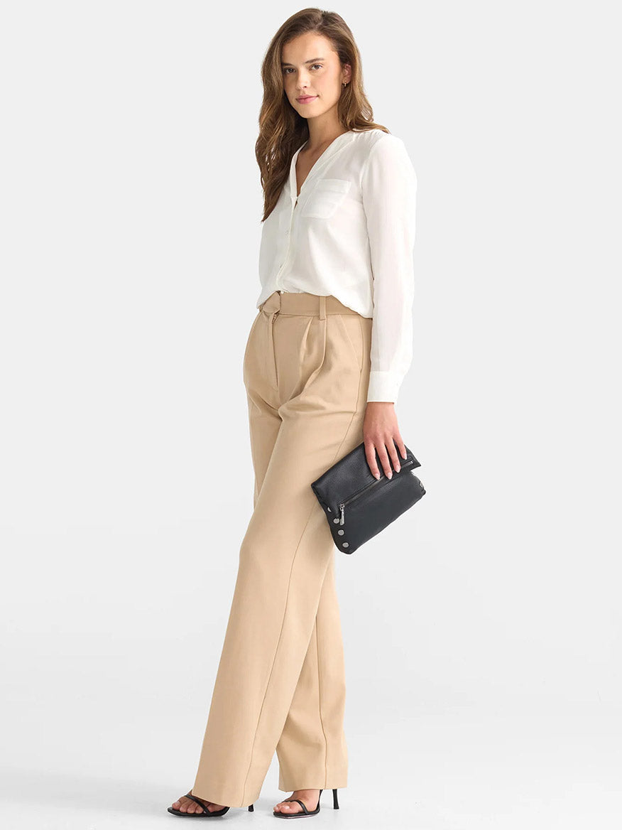 A person stands wearing a white blouse and beige trousers, holding the Hammitt Los Angeles VIP Medium Clutch in Black with an adjustable crossbody strap.