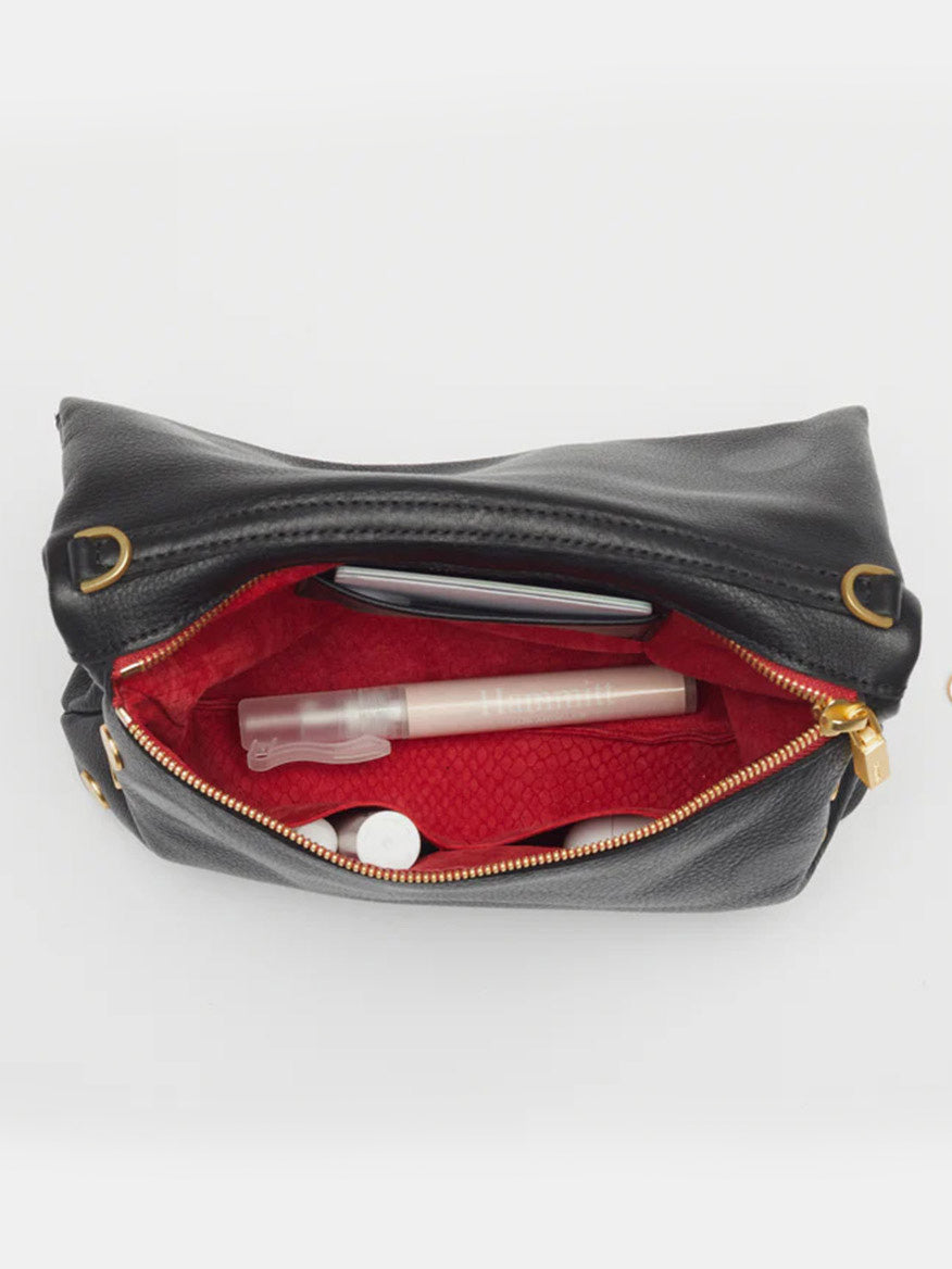 An open Hammitt Los Angeles VIP Medium Clutch in Black crafted from buttery black leather with brushed gold hardware. Its red interior reveals a small spray bottle, white containers, and a pink item. The zipper is partially unzipped, offering a glimpse of the contents inside.