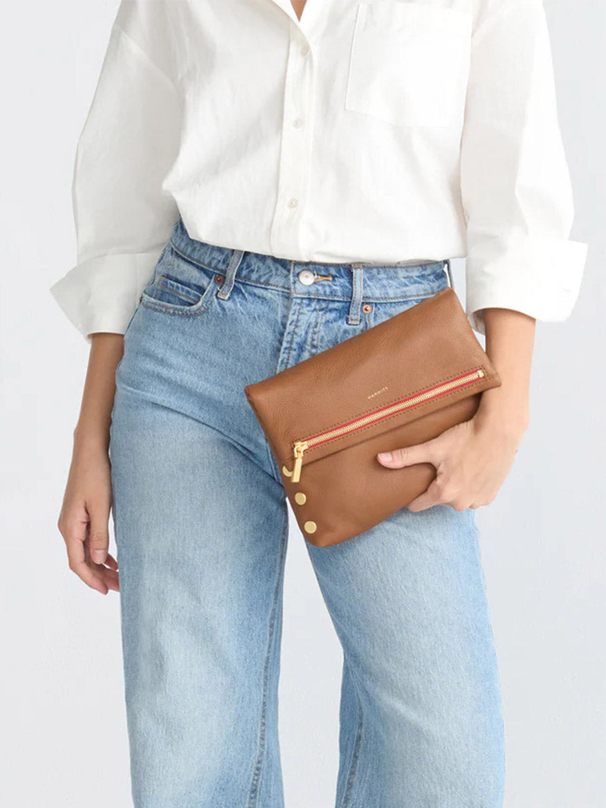 A person in a white shirt and blue jeans holds a Hammitt Los Angeles VIP Medium Clutch in Mahogany Pebble, featuring brushed gold rivets and a red zipper.