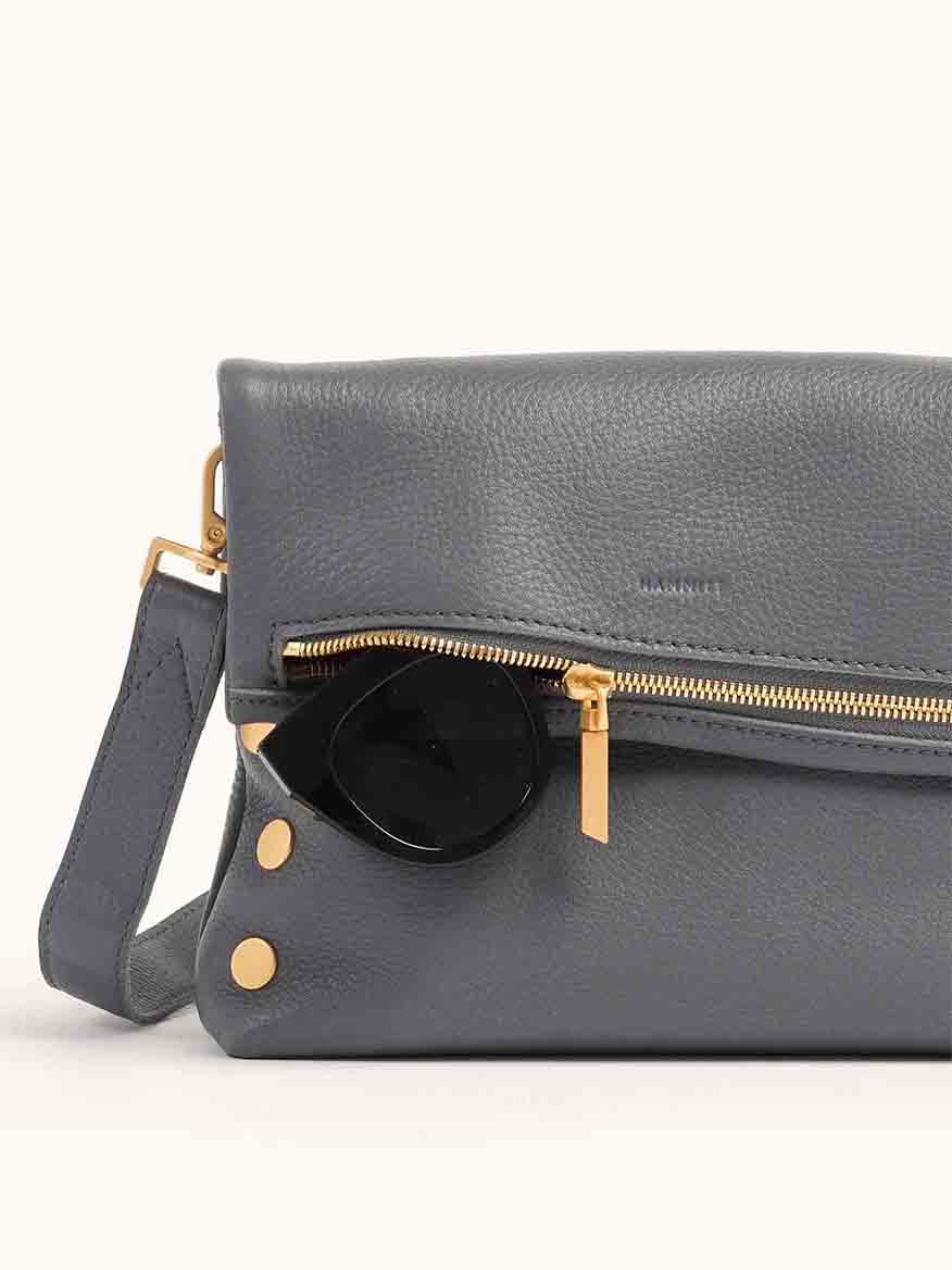 This Hammitt Los Angeles VIP Medium Clutch in Sonnet Grey boasts pebbled leather with a gold zipper and hardware. It has a small side strap and is perfectly complemented by black sunglasses partially tucked into the zipper.