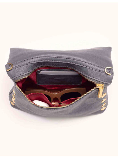 A partially open Hammitt Los Angeles VIP Medium Clutch in Sonnet Grey, crafted from pebbled leather, holds a wallet, glasses, and a small white item against a plain background.