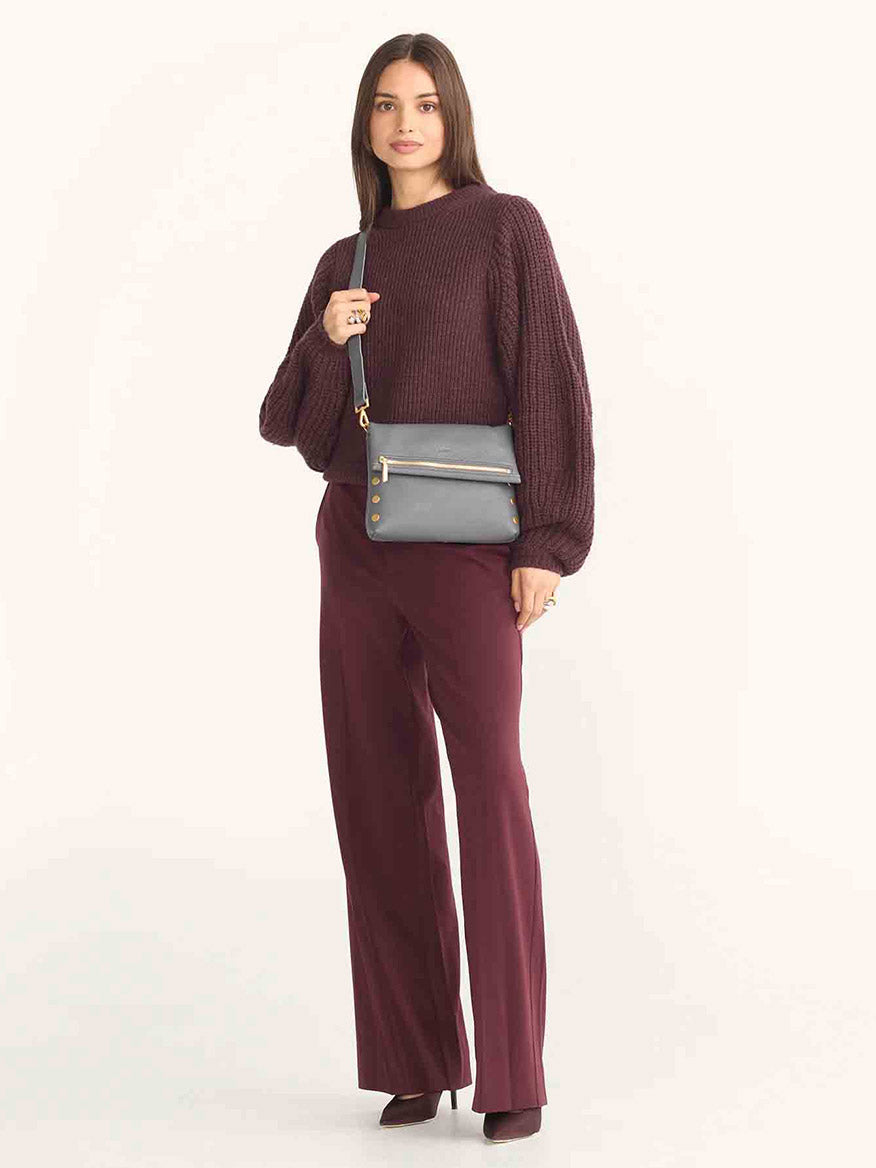 A woman in a maroon sweater and pants elegantly holds a Hammitt Los Angeles VIP Medium Clutch in Sonnet Grey, crafted from luxurious pebbled leather, against a plain backdrop.