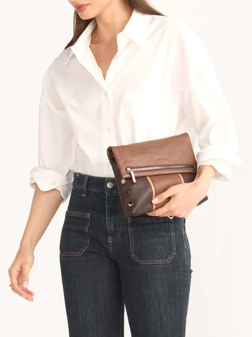 A person in a white shirt and dark jeans is holding the Hammitt Los Angeles VIP Medium Clutch in Orchestra Brown, which features an exterior phone pocket.