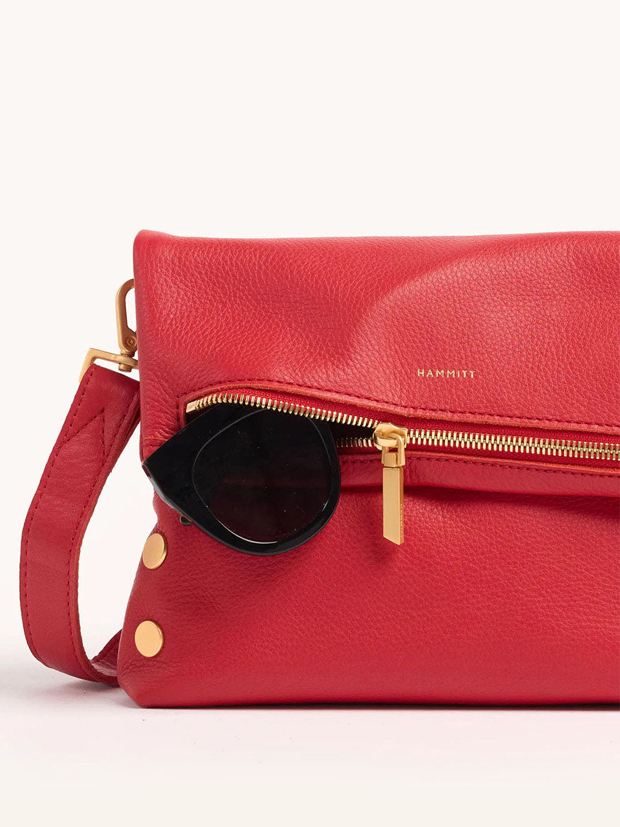 The Hammitt Los Angeles VIP Medium Clutch in Poppy Red leather, featuring a gold zipper and accents, effortlessly transforms into a chic crossbody bag with round black sunglasses partially tucked behind the flap.