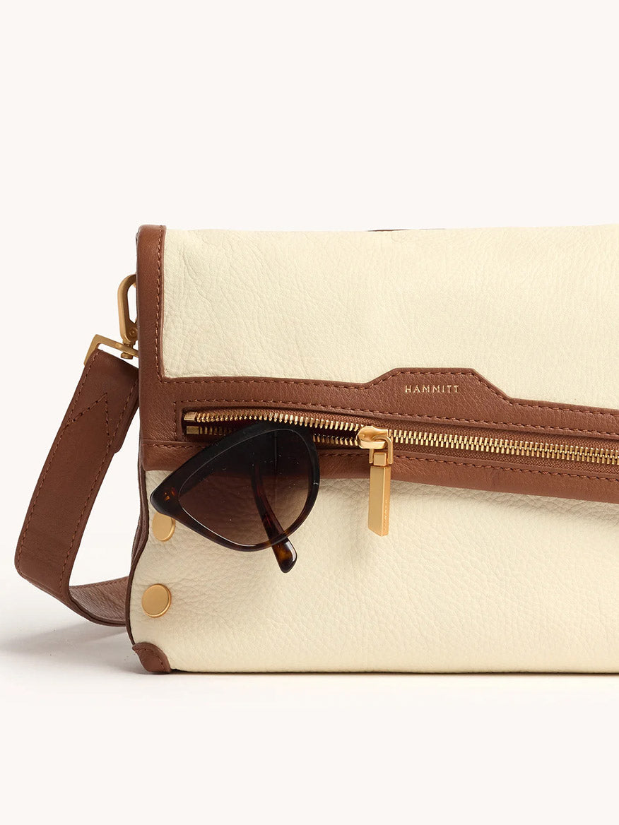 The Hammitt Los Angeles VIP Medium Clutch in Courtyard Cream is a cream and brown leather bag with a gold zipper, gunmetal rivets, and dark sunglasses hooked on its front pocket.