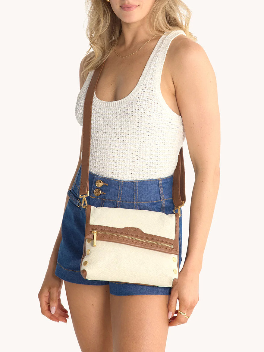 A woman wearing a white knit tank top and blue high-waisted shorts holds the Hammitt Los Angeles VIP Medium Clutch in Courtyard Cream, featuring elegant gold hardware.