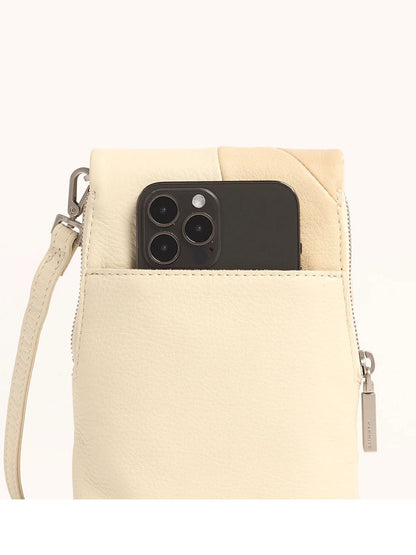 A smartphone peeks out of a light beige Leather crossbody bag, the Hammitt Los Angeles VIP Mobile in Coastal Cream, showcasing a wrist strap and chic metal zipper pull.