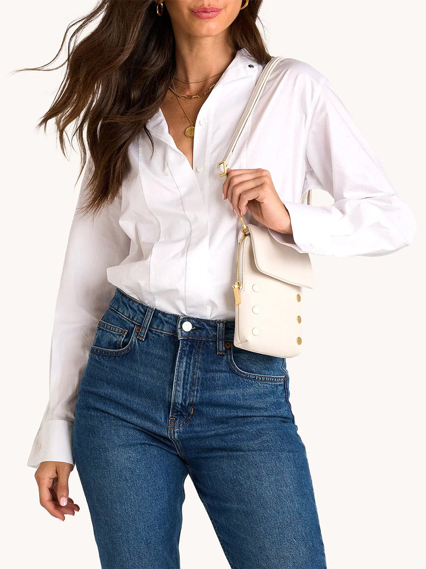 Woman wearing a white button-down shirt and blue jeans, carrying the Hammitt Los Angeles VIP Mobile in Calla Lily White/Brushed Gold, a small crossbody bag with gold accents; shown from shoulders to hips.