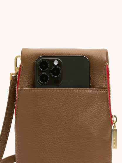 A black smartphone is partially tucked into the cellphone pocket of the Hammitt Los Angeles VIP Mobile in Mahogany Pebble with brushed gold accents and a red zipper, carried crossbody for a chic look.