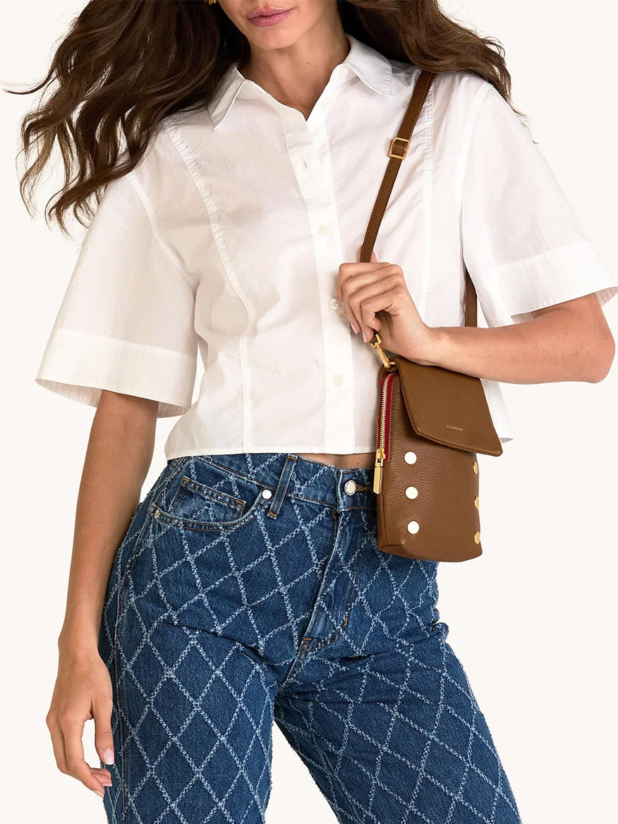 Woman wearing a white short-sleeve button-up shirt, patterned blue jeans, and carrying the Hammitt Los Angeles VIP Mobile in Mahogany Pebble/Brushed Gold/Red Zip crossbody bag with gold accents and a cellphone pocket.