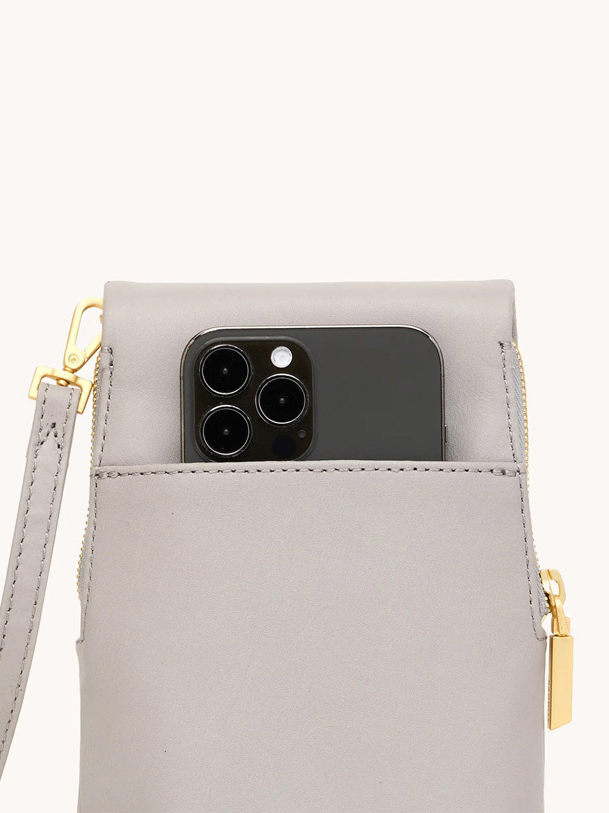 A close-up of the Hammitt Los Angeles VIP Mobile in Stormy/Brushed Gold, a gray pebble leather crossbody bag with a gold zipper and strap, holding a black smartphone with triple lenses in its front pocket.