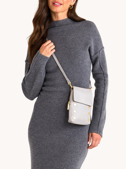 A woman wearing a gray knit dress holds the Hammitt Los Angeles VIP Mobile in Stormy/Brushed Gold, a light gray pebble leather crossbody bag with gold hardware.