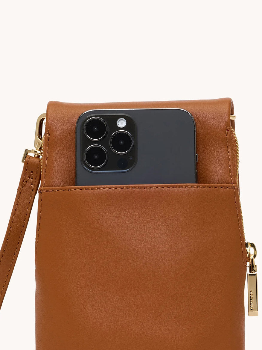 A black smartphone, possibly a VIP Mobile, is partially tucked into the front pocket of a Hammitt Los Angeles VIP Mobile bag in Cocoa Raffia/Gold with a gold zipper and wrist strap.