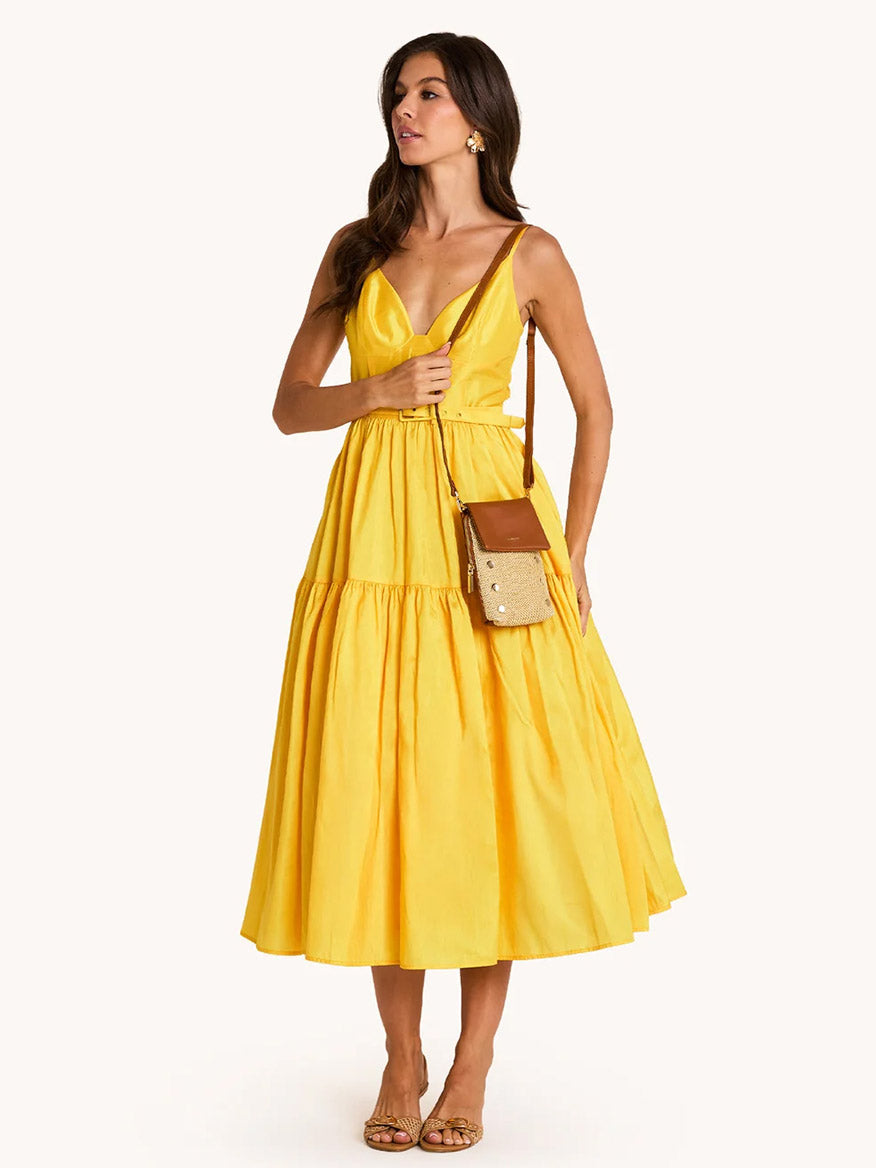 Woman wearing a sleeveless yellow midi dress, brown sandals, and carrying the Hammitt Los Angeles VIP Mobile in Cocoa Raffia/Gold crossbody bag, standing against a plain light background.