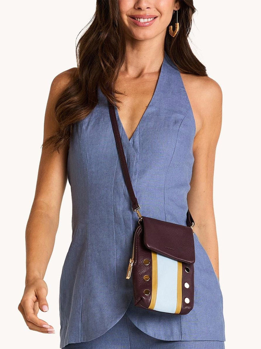 A woman in a sleeveless blue halter jumpsuit carries the Hammitt Los Angeles VIP Mobile crossbody bag in Regatta Stripe/Gold, featuring maroon, white, light blue color blocks, and metallic studs.