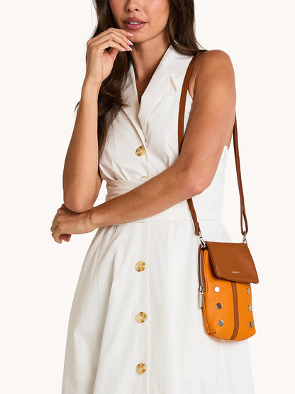 A woman wears a sleeveless white button-down dress and carries the Hammitt Los Angeles VIP Mobile in Tailored Tangerine/Silver, a chic small orange crossbody bag with silver studs.