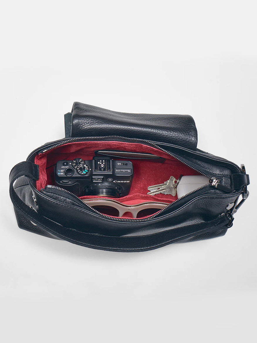 Top view of the open Hammitt Los Angeles VIP Satchel in Black/Gunmetal, showing items inside, including a camera, a pair of sunglasses, keys, and another smaller black item on a red interior lining. The VIP Satchel comes with a removable crossbody strap for versatile styling.