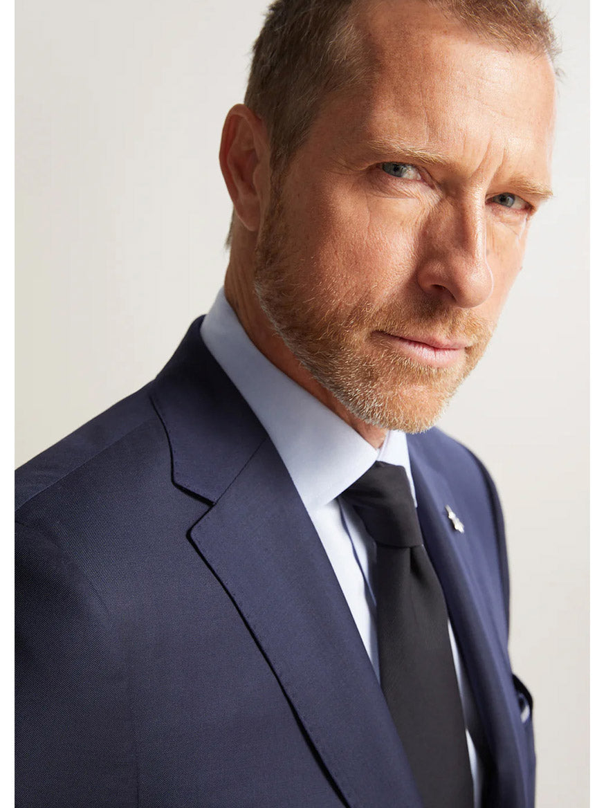 A man with a beard, wearing the Hickey Freeman (Heritage Gold) Navy Traveler Honey Way Blazer over a light blue shirt and black tie, exudes the aura of a modern gentleman as he glances confidently towards the camera.