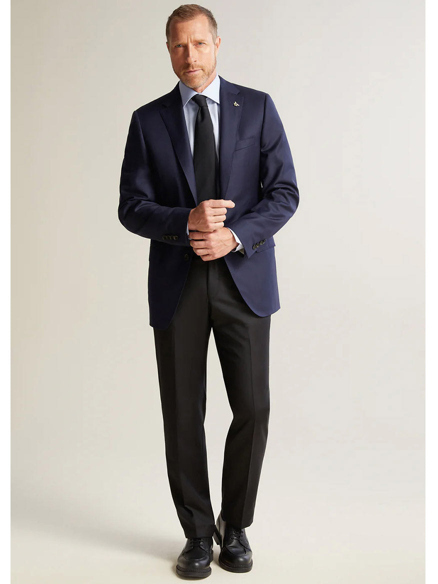 A modern gentleman in a dark suit and tie stands against a plain background, adjusting his sleeves with the finesse of someone wearing the Hickey Freeman (Heritage Gold) Navy Traveler Honey Way Blazer.