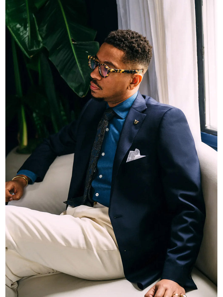 A modern gentleman in a Hickey Freeman (Heritage Gold) Navy Traveler Honey Way Blazer and glasses sits on a white couch near a large plant and window.