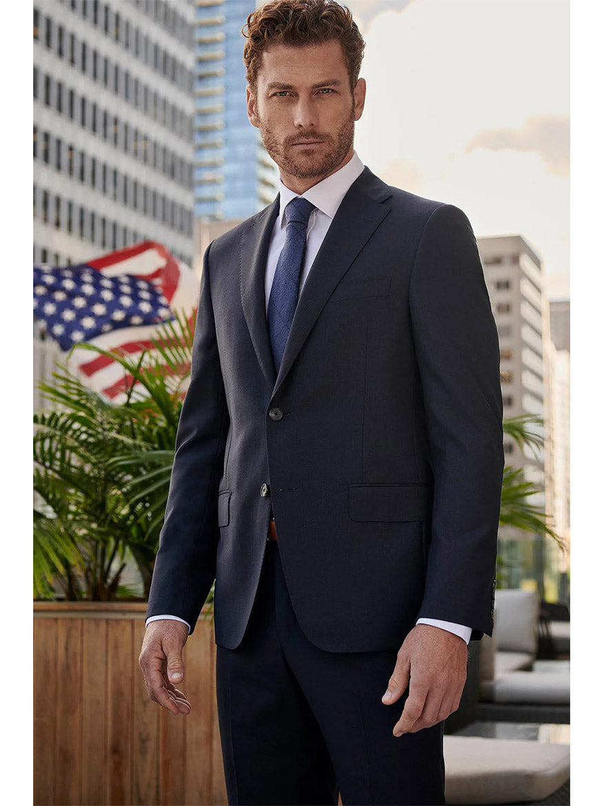 A man in a sophisticated Hickey Freeman (Heritage Gold) Navy Sharkskin Infinity Suit stands outdoors with an American flag and buildings in the background, showcasing elegance crafted from Super 140s wool by the renowned Carlo Barbera mill.