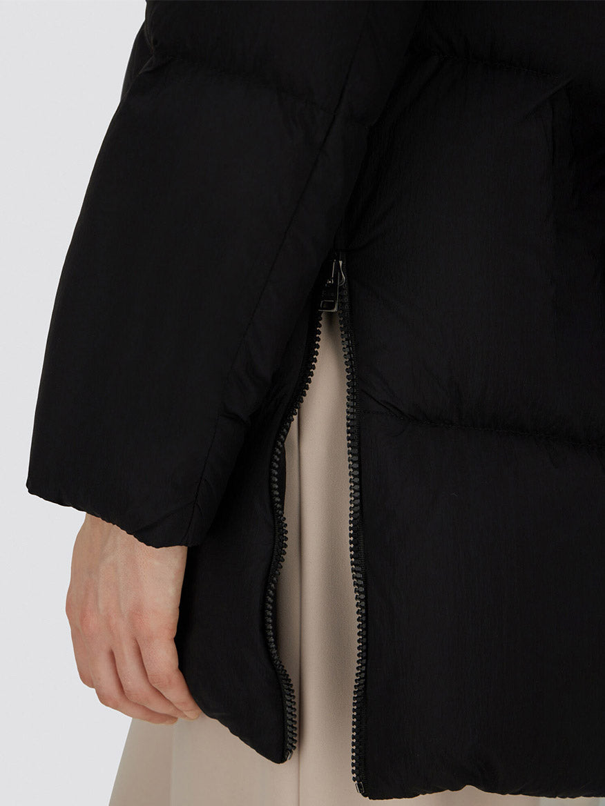 A close-up of a hand next to the side zipper of the Herno A-Shape Jacket in Ecoage in Black, made from recycled nylon with goose down padding, with a beige garment partially visible underneath.