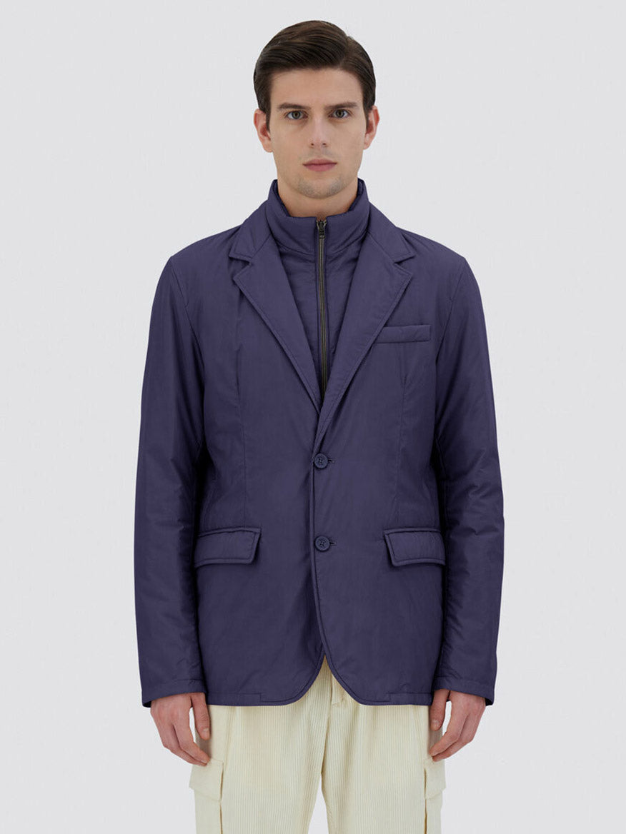 A person stands facing forward with a neutral expression, wearing the Herno Blazer in Ecoage in New Blue over a padded jacket made of recycled nylon microfibre. The background is plain light gray.