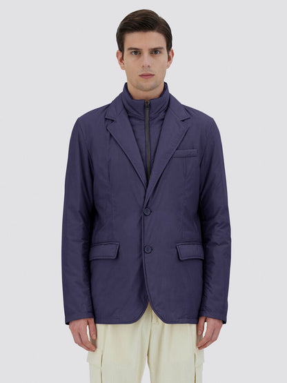 A person stands facing forward with a neutral expression, wearing the Herno Blazer in Ecoage in New Blue over a padded jacket made of recycled nylon microfibre. The background is plain light gray.