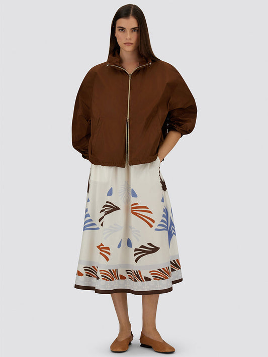 A woman wears a brown zip-up jacket, brown flats, and the Herno Summer Print Cotton Skirt in Multicolor—white with vibrant abstract patterns—standing against a plain background.