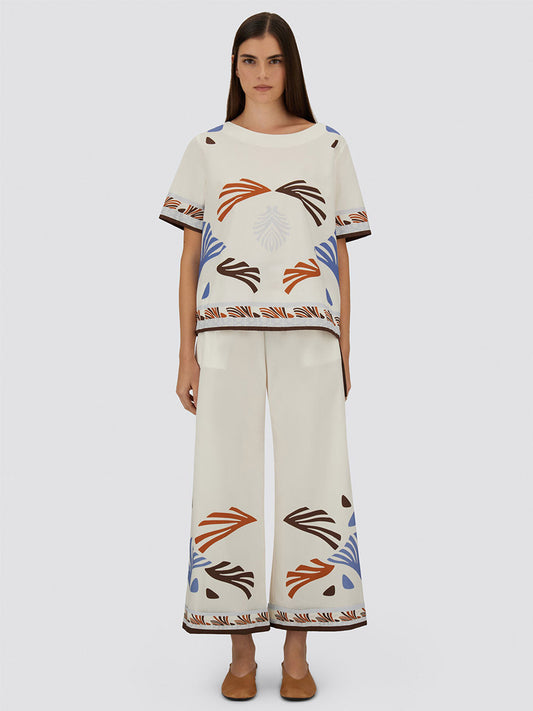A woman faces forward in the Herno Summer Print Cotton Top in Multicolor and cream wide-leg pants with orange, blue, and brown geometric patterns, paired with brown shoes. The Italian-made ensemble is set against a plain light background.