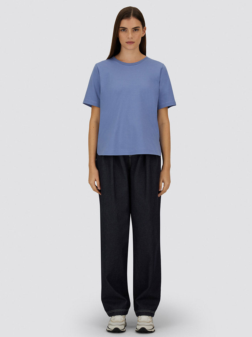 A person stands facing forward wearing a Herno Superfine Cotton Stretch T-Shirt in light blue, loose dark jeans, and white sneakers against a plain light gray background.