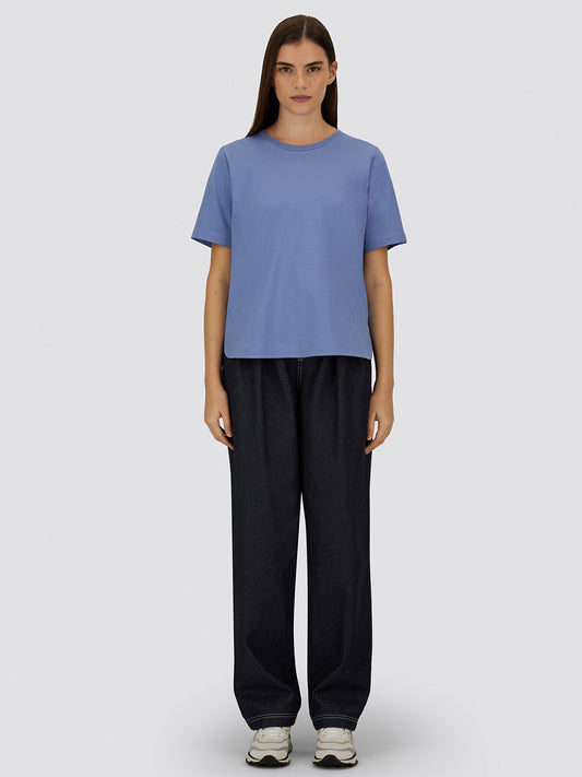 A person stands facing forward wearing a Herno Superfine Cotton Stretch T-Shirt in light blue, loose dark jeans, and white sneakers against a plain light gray background.