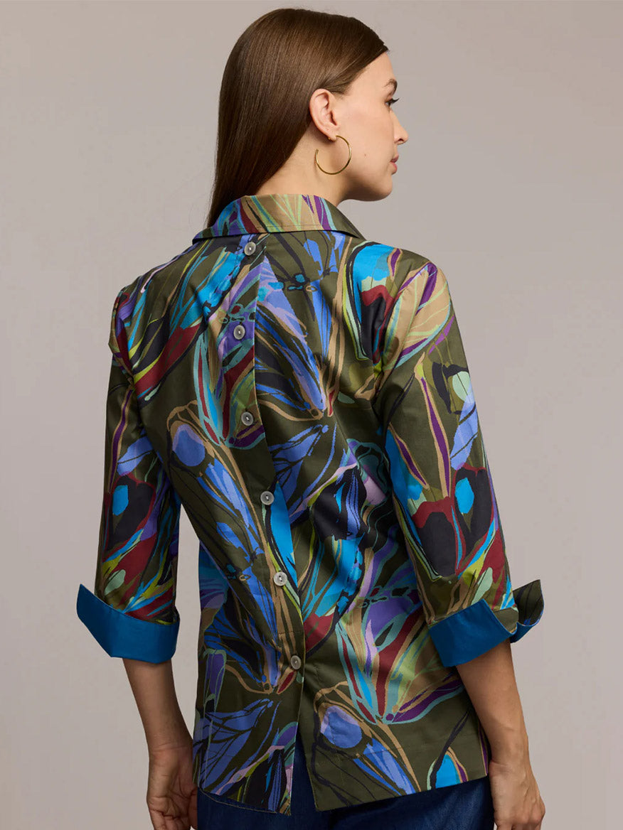 A woman faces away, wearing the Hinson Wu Aileen 3/4 Sleeve Abstract Camo Shirt with sapphire accents and button-back detail.