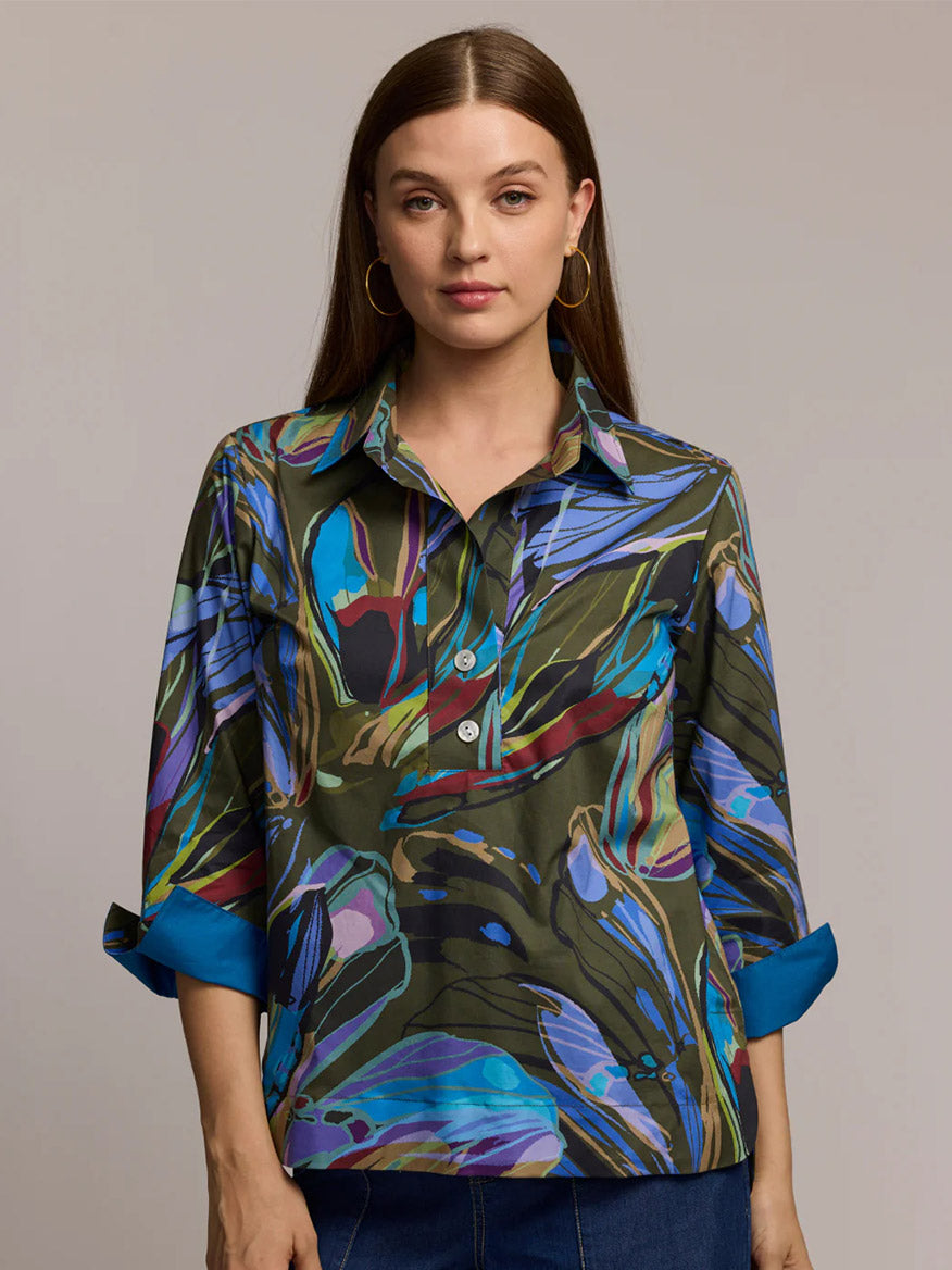 A woman stands against a plain background, wearing the Hinson Wu Aileen 3/4 Sleeve Abstract Camo Shirt With Sapphire cuffs and gold hoop earrings.