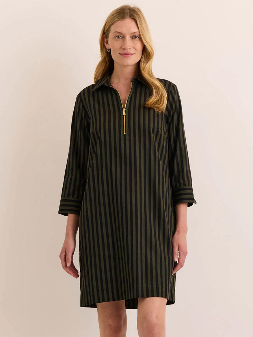 A woman stands against a plain beige background wearing the Hinson Wu Alessa 3/4 Sleeve Classic Stripe Dress in Oregano/Black, which features black and olive-green vertical stripes, three-quarter sleeves, a front gold zipper detail, and is crafted from premium cotton fabric.