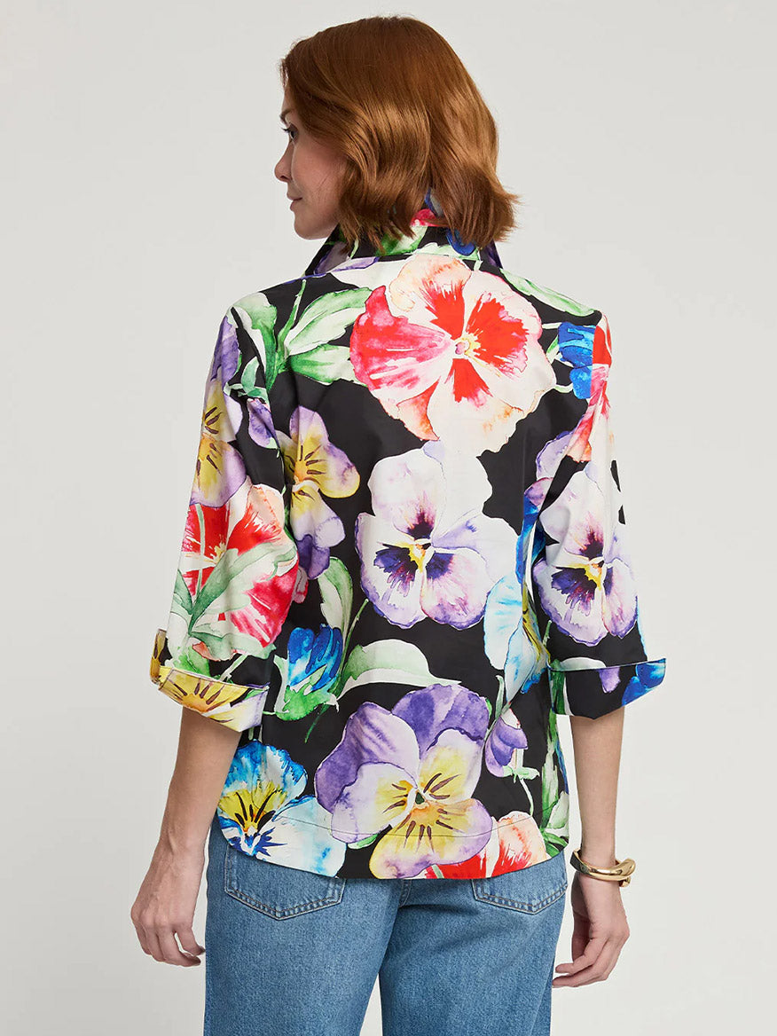 A person with short, brown hair is seen from behind, wearing the Hinson Wu Charlotte 3/4 Sleeve Pansy Print Top. The black blouse with a colorful Pansy Print has its 3/4 sleeves rolled up, giving it a popover style look. Paired with blue jeans and a gold bracelet, the outfit is effortlessly chic.