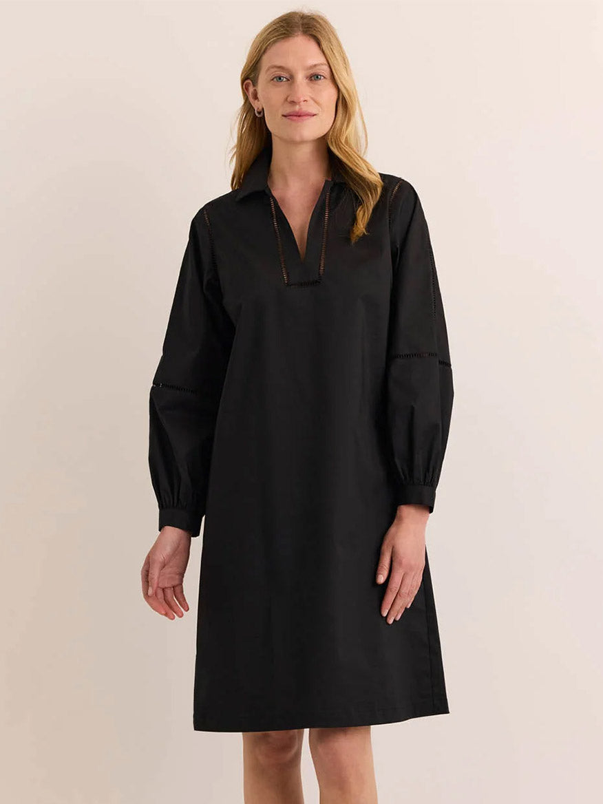 A fair-skinned woman with blonde hair wears the Hinson Wu Laura Long Sleeve Lace Trim Dress in Black—a knee-length dress featuring a V-neckline, collar, long sleeves, and subtle ladder lace detailing. Crafted from stretch polished cotton for effortless comfort and a relaxed fit, the dress combines elegance with ease. She stands against a plain, off-white background.