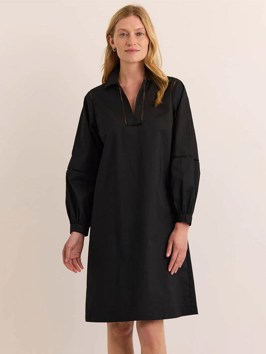 A fair-skinned woman with blonde hair wears the Hinson Wu Laura Long Sleeve Lace Trim Dress in Black—a knee-length dress featuring a V-neckline, collar, long sleeves, and subtle ladder lace detailing. Crafted from stretch polished cotton for effortless comfort and a relaxed fit, the dress combines elegance with ease. She stands against a plain, off-white background.