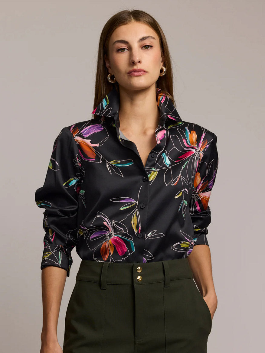 A woman stands with hands in pockets, wearing the Hinson Wu Margot Long Sleeve Floral Stencil Shirt in Black—a relaxed fit, cotton blend satin button-up with a colorful floral print—paired with olive green pants against a plain background.