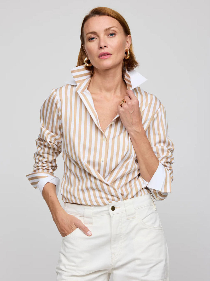 Wearing the Hinson Wu Margot Long Sleeve Stripe Shirt in Latte/White with a popped collar, a woman with shoulder-length hair showcases timeless style. Her white pants and hoop earrings complete the polished look as she stands against a plain background, one hand in her pocket.