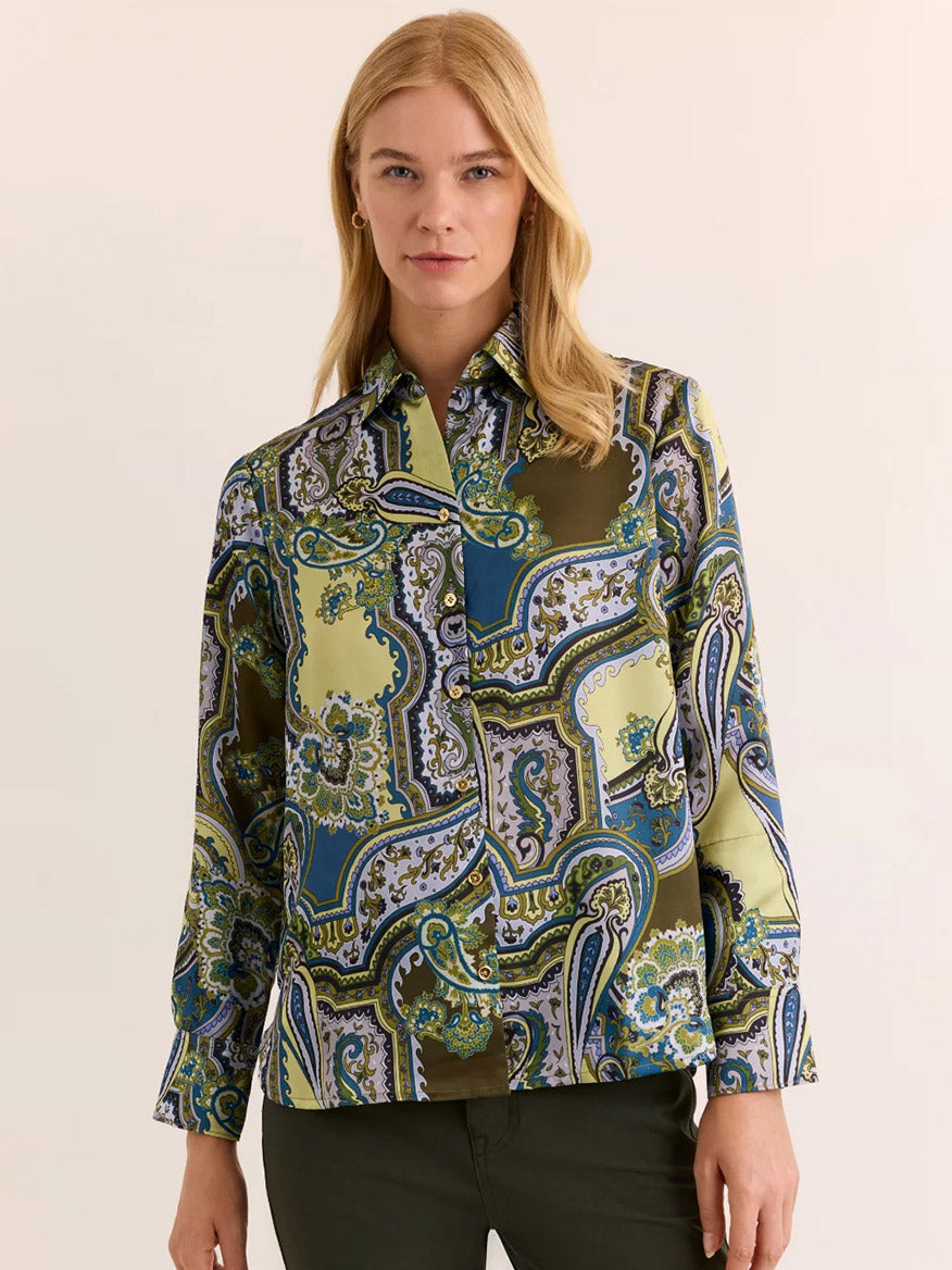 A woman stands facing forward against a plain background, dressed in the Hinson Wu Margot Long Sleeve Opulent Paisley Shirt in Olive Paisley, a button-up shirt crafted from luxurious Italian polyester with a vibrant multicolored paisley pattern.